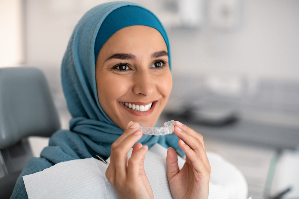 Woman in a hijab in a dental office smiling while holding a clear aligner tray