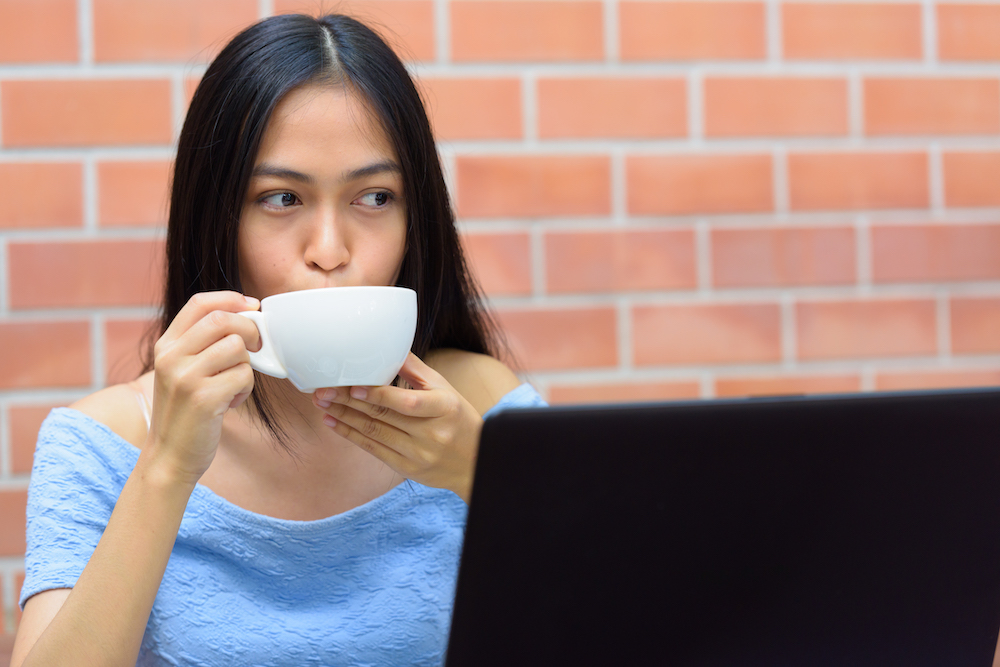 Asian teenage girl drinking coffee while thinking with laptop against brick wall